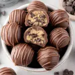 A plate of chocolate-covered Cookie Dough Truffles with a bite taken out showing the soft dough center