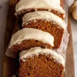 A sliced loaf of moist pumpkin bread with cream cheese frosting on a wooden board
